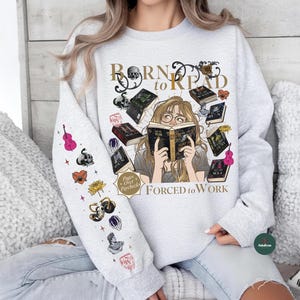 May include: A light gray sweatshirt with the text "Born to Read" and "Forced to Work". The graphic features a woman reading surrounded by books. The left sleeve has various colorful illustrations including a pink violin and a skull.