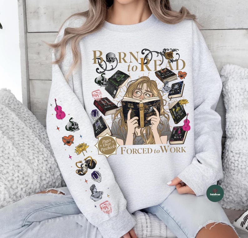 May include: A light gray sweatshirt with the text "Born to Read" and "Forced to Work". The graphic features a woman reading surrounded by books. The left sleeve has various colorful illustrations including a pink violin and a skull.
