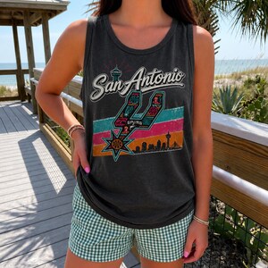 Puede incluir: Una camiseta sin mangas de color gris carb&oacute;n con un estampado gr&aacute;fico que dice San Antonio, un logotipo estilizado de los Spurs, un fondo de rayas coloridas y la silueta de una ciudad. La camiseta se combina con pantalones cortos de cuadros vichy.