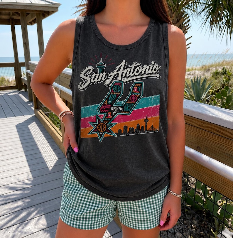 Puede incluir: Una camiseta sin mangas de color gris carb&oacute;n con un estampado gr&aacute;fico que dice San Antonio, un logotipo estilizado de los Spurs, un fondo de rayas coloridas y la silueta de una ciudad. La camiseta se combina con pantalones cortos de cuadros vichy.