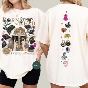 May include: Cream-colored t-shirt with the text "BORN to READ" and "FORCED to WORK". The front features a graphic of a person reading surrounded by books and skulls. The back has a vertical design with various illustrations.