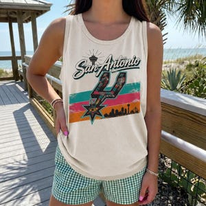 Puede incluir: Una camiseta sin mangas de color crema con un estampado gr&aacute;fico que dice San Antonio en letra cursiva sobre un logotipo de espuela estilizado, rayas horizontales coloridas y una silueta negra del horizonte de la ciudad con la torre.