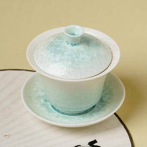 Könnte beinhalten: Ein Gaiwan-Teeservice aus Keramik, bestehend aus einer Schale, einem Deckel mit kleinem Knauf und einer Untertasse. Das Set weist eine hellminzgrüne und weiße kristalline Glasur auf, die eine zarte, frostähnliche Textur auf der Oberfläche erzeugt.