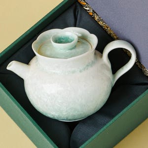 May include: A round ceramic teapot with a pale mint green and white crystalline glaze, featuring a scalloped rim, a matching lid with a small knob handle, and a curved spout, presented inside a dark green fabric-lined gift box.