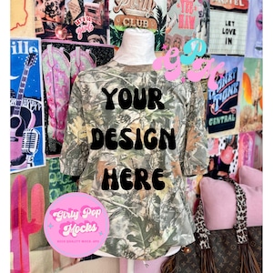 May include: A short-sleeved camouflage t-shirt with the text "YOUR DESIGN HERE" in black. The shirt is displayed on a mannequin against a backdrop of colorful posters and a pink pillow. A pink and white logo that says "Girly Pop Mocks" is in the lower left corner.
