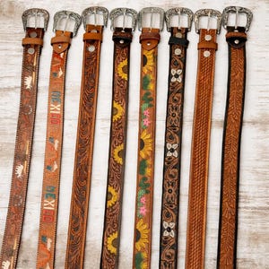 May include: Eight leather belts arranged vertically, each featuring a silver-toned metal buckle. The belts display various embossed patterns, including floral motifs, sunflowers, cacti, geometric textures, and the word MEXICO with pyramid icons.