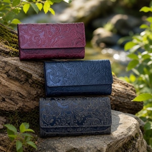 May include: Three rectangular wallets featuring an embossed floral pattern on the exterior. The top wallet is deep red, the middle wallet is navy blue, and the bottom wallet is a dark charcoal gray, each with a flap closure and a structured design.