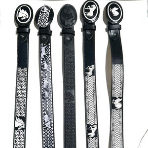 May include: Five black leather belts arranged vertically, each featuring a distinct oval buckle. The belts display intricate white embroidered patterns, including geometric shapes, horse motifs, and repeating Greek key designs, with matching oval buckles.