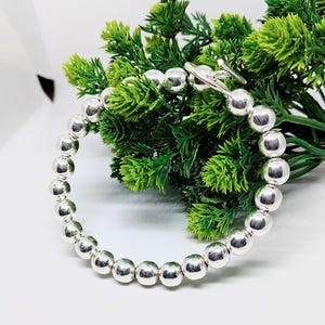 Pode incluir: Uma pulseira de contas de prata com fecho de alternância. A pulseira é composta por contas redondas e polidas de prata. A pulseira é exibida em uma superfície branca com folhagem verde ao fundo.