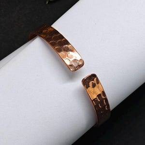 Può includere: Bracciale aperto in rame con texture martellata. Il bracciale è di un caldo color marrone-rossiccio e ha un design semplice e aperto. Il bracciale è mostrato su una superficie bianca con sfondo nero.