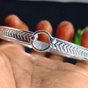 May include: A silver-colored bracelet with a geometric arrow pattern. The bracelet has a circular clasp and is held in a hand. The design features a series of repeating arrow shapes along the band.