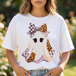 May include: A white short-sleeve t-shirt featuring a central graphic of a ghost wearing a large bow. The ghost has black oval eyes and eyelashes, with sections of the body and bow decorated in purple checkered and orange leopard print patterns.