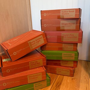 May include: A collection of rectangular KiwiCo Labs project boxes in orange, red, and green. The boxes are labeled with project names including Rubber Band Blasters, Mega Hand, Rapid-Fire Disc Launcher, Hydraulic Claw, Staircase Automaton, Bowling Arcade, Transforming Tops, Party Animal Disco Light, Pop-Up Pencil Box, Mechanical Grabber, and Wrist Rockets.
