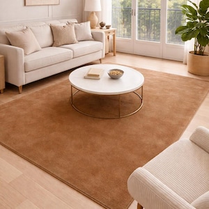 May include: A large, rectangular area rug in a solid, warm tan color with a soft, plush texture. The rug features a clean, finished edge and is positioned centrally on a light-colored wooden floor, providing a neutral foundation for the room.