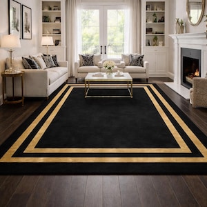 May include: A black rectangular rug with a gold border, placed in a living room. The rug sits on a dark wood floor, with white sofas and a fireplace visible. The room is illuminated by natural light.