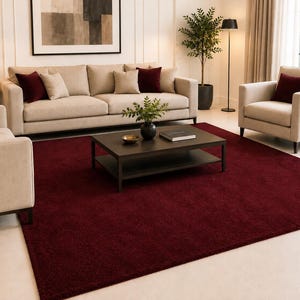 May include: A large, rectangular area rug in a deep, solid burgundy color with a plush, textured pile. The rug is positioned centrally on a light-colored floor, serving as a foundation for a living room furniture set including a sofa and chairs.