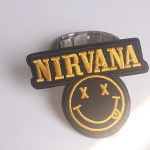 May include: Embroidered patch with the word "NIRVANA" in gold thread on a black rectangular background. Below, a circular patch with a black background displays a yellow smiley face with crossed-out eyes and a protruding tongue.