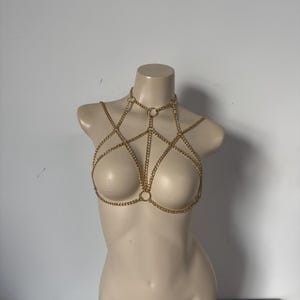 May include: A gold-toned metal body chain harness featuring a series of interconnected curb chains and circular metal rings. The design includes a choker-style neck piece that extends into a geometric web pattern covering the chest area.