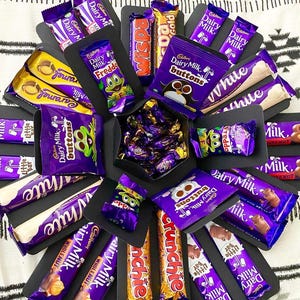 Puede incluir: Una selección de barras de chocolate y caramelos Cadbury, incluyendo Dairy Milk, Freddo, Wispa y Crunchie, dispuestos en un círculo. El embalaje presenta principalmente colores púrpura, blanco y dorado. Se ven varios productos de chocolate.