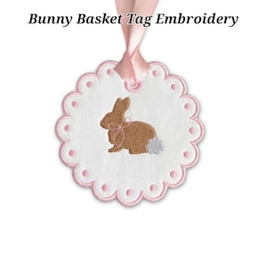 May include: White, scalloped-edge tag with pink trim and a brown embroidered bunny wearing a pink bow. The tag is suspended by a pink ribbon. Text at the top reads "Bunny Basket Tag Embroidery".