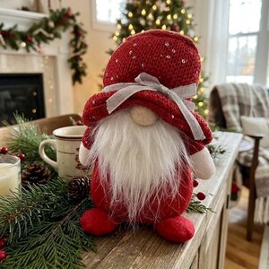 May include: A plush gnome figurine featuring a red knitted hat with subtle silver sequins and a silver ribbon bow. The gnome has a long, white faux fur beard, a round beige nose, and red fabric feet. The body is round and covered in red fabric.