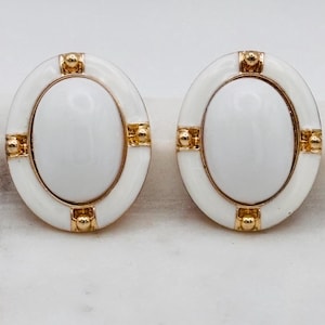 May include: A pair of oval-shaped earrings. Each earring features a white enamel center surrounded by a gold-tone frame and white enamel border. Gold-tone accents are placed at the top, bottom, and sides of the oval shape. The earrings are set against a white background.