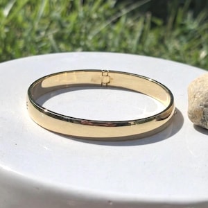 May include: A polished gold-tone metal bangle bracelet with a smooth, reflective surface. The circular bracelet features a visible hinged clasp mechanism on one side, allowing it to open and close securely around the wrist for a comfortable fit.