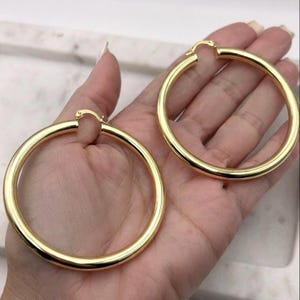 May include: A pair of large, polished gold-tone hoop earrings with a smooth, tubular design. Each earring features a classic circular shape with a hinged post closure mechanism at the top, showcasing a bright, reflective metallic finish throughout.