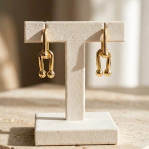 May include: A pair of gold-toned earrings featuring a U-shaped link design with rounded ball ends. The earrings are displayed on a T-shaped stand. Each earring consists of a vertical bar connected to a curved base with two small spheres at the bottom.