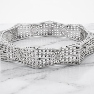 May include: A silver-toned hexagonal bangle bracelet featuring an intricate openwork lattice design. The structure is composed of geometric segments with circular accents at each joint, creating a repeating pattern throughout the entire piece.