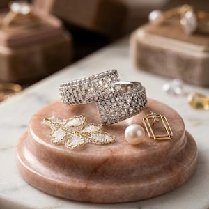 May include: Two silver rings encrusted with small, sparkling stones are displayed on a pink marble pedestal. A gold-trimmed, white lace leaf, a pearl, and a gold earring with a small diamond are also present.