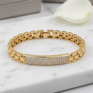 May include: Gold-toned bracelet with a rectangular center section encrusted with small, sparkling stones. The bracelet has a linked design and a secure clasp. The jewelry is displayed on a white marble surface.