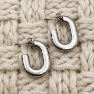 May include: A pair of silver-colored, U-shaped hoop earrings. The earrings have a smooth, polished finish and a post-and-clasp closure. The earrings are displayed on a cream-colored, textured surface.
