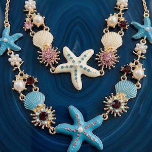 May include: A gold-toned necklace featuring starfish, seashells, and floral accents. The starfish are light blue and white, adorned with pink and blue crystals. The necklace includes faux pearls and red and pink gemstones, creating a nautical theme.