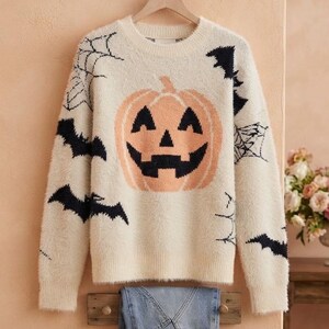 May include: A cream-colored fuzzy long-sleeve sweater featuring a large orange jack-o'-lantern graphic on the center chest, surrounded by black bat silhouettes and black spiderweb patterns printed across the front and sleeves.