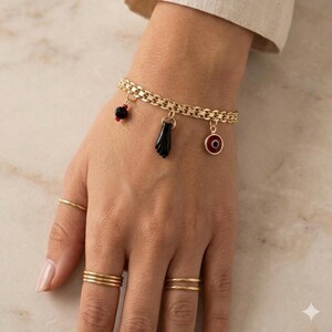 May include: Gold-toned bracelet with three charms: a black hand, a red and black bead, and a red and black eye. The bracelet is worn with several gold rings. The jewelry is displayed on a hand.