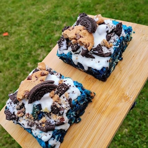 May include: Two square, blue-colored dessert bars topped with white frosting, cookie pieces, and chocolate sandwich cookies. The desserts sit on a light brown wooden board against a green grassy background.
