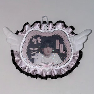 May include: A decorative heart-shaped item featuring a photo of a person. The item has a pink and black polka-dot border with white wings and a small bow. A silver chain is attached for hanging.