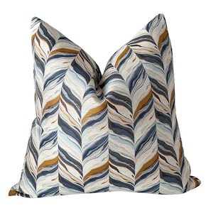 May include: Decorative throw pillow with a repeating pattern of abstract, flowing lines in shades of blue, tan, and cream. The pillow has a soft, textured appearance and a neutral background, suitable for home decor.