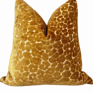 May include: A square throw pillow featuring a mustard yellow velvet fabric with a raised, textured animal-inspired spot pattern in a lighter cream color. The pillow has a soft, plush appearance with a distinct indentation at the top center.