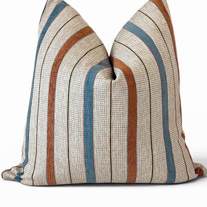 May include: A square throw pillow featuring a textured cream-colored base with vertical stripes in shades of rust orange, muted blue, and thin dark brown lines. The fabric has a visible woven grid pattern throughout the entire surface of the pillow.