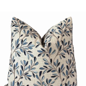 May include: Decorative throw pillow with a beige linen-like fabric, embroidered with a pattern of blue leaves and vines. The pillow has a soft, inviting appearance, suitable for home decor.