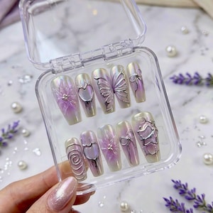 May include: A set of iridescent, long, rectangular press-on nails in a clear plastic case. Each nail features a design, including a flower, heart, butterfly, and bow, with silver-colored metal accents. The nails have a pearlescent finish with lavender and white hues.