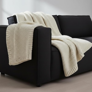 May include: A cream-colored knitted throw blanket draped over a black sofa. The blanket has a textured, raised pattern and a ribbed border. The sofa has a modern, minimalist design.