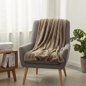 Pode incluir: Uma manta bege texturizada sobre uma poltrona cinza com pernas de madeira. A manta tem um padrão tecido e é exibida em uma sala bem iluminada com uma planta e livros em uma mesa lateral.