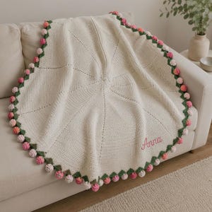 Könnte beinhalten: Eine runde, cremefarbene Häkeldecke mit einem strukturierten, strahlenförmigen Stichmuster. Der Rand ist mit einer dunkelgrünen, gewellten Borte versehen, die mit kleinen, mehrfarbigen rosa und weißen Pompons verziert ist. Der Name Anna ist in rosa Schreibschrift eingestickt.