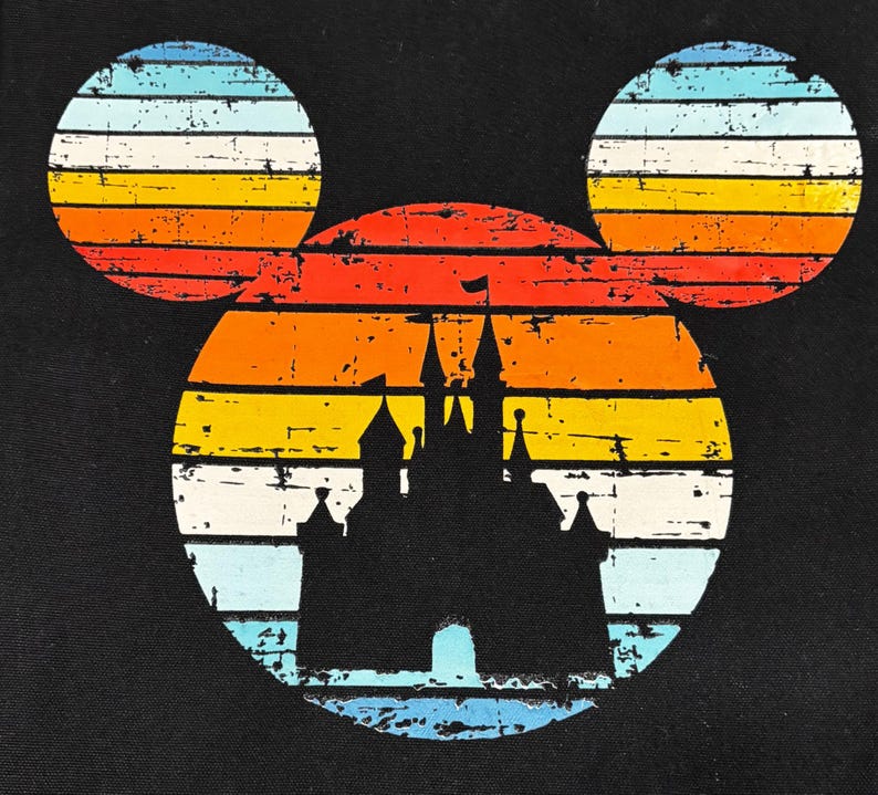 May include: Black graphic featuring a silhouette of a castle within a Mickey Mouse head shape. The design has a retro sunset color scheme of orange, yellow, and blue stripes, with a distressed, vintage look.