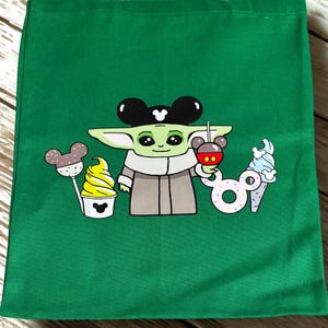 May include: Green tote bag featuring a cartoon character with large ears, holding a Mickey Mouse-shaped drink. The design includes a cupcake, a lollipop, and an ice cream cone, all with Mickey Mouse ears.