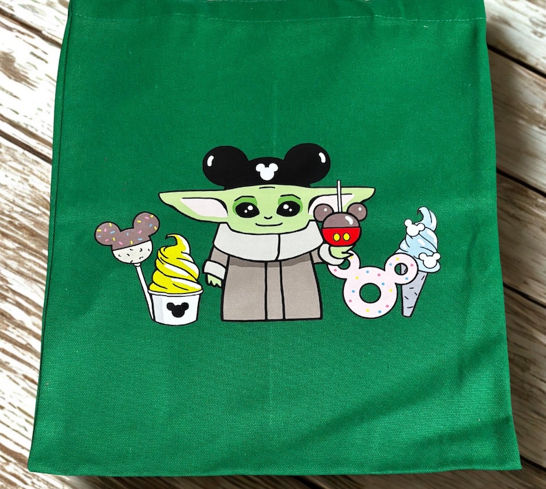 May include: Green tote bag featuring a cartoon character with large ears, holding a Mickey Mouse-shaped drink. The design includes a cupcake, a lollipop, and an ice cream cone, all with Mickey Mouse ears.