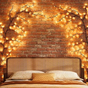 May include: A bedroom scene featuring a brick wall adorned with two decorative branches lit with warm, glowing fairy lights. A bed with a woven headboard and pillows is in front of the wall. A nightstand and a clock are visible.